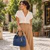 A woman stands on a cobblestone street in a sunny outdoor setting, wearing a white short-sleeve button-up shirt, tan wide-leg trousers, and holding the dark blue woven leather bag with gold hardware.